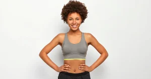 Smiling woman in a grey sports bra with hands on hips, showing a toned midsection after weight loss.