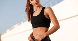 Woman in a black sports bra outdoors adjusting her leggings, showing a flat, defined midsection.